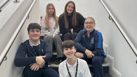 The photo shows a group of five people sitting together on an indoor staircase. Three individuals are seated on the steps toward the back, and two are positioned lower down. The staircase has smooth white walls on both sides with metal handrails running along the length of the stairs. Some of the group are wearing clothing with school emblems, including dark jumpers, sports-style tops, and a white polo shirt