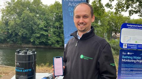 Tom pictured looking into the camera and smiling, holding up a smartphone with a screen showing two graphs. He's standing in front of a trestle table which has a large black barrel on it that reads (upside down) 'alert system' in blue lettering. Behind that is the River Severn, lined by trees on the bank and with a slipway leading down to it. Alex is wearing a black fleece with a green logo on it reading 'Environment Agency.'
