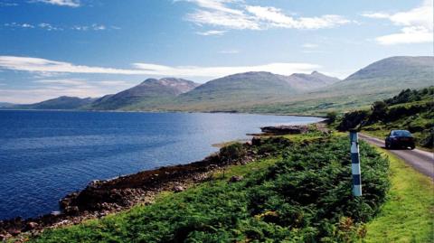 A car travels down a road, by the sea, on the Isle of Mull. The car is heading towards some mountains. 