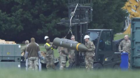 Image of an air base, with a forklift truck in the background. In the foreground can be seen a number of military personnel in khaki uniforms who are guiding explosive ordnance into position.