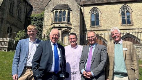 A mixed group of formally dressed men stand in front of a historic stone building.