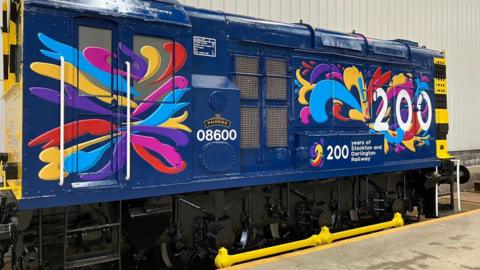 A dark blue shunting engine with colourful splashes of paint and a big "200" in white letters, beneath which it reads "years of Stockton and Darlington Railway". The engine is rectangular shaped and the four wheels beneath it are shining black with bright yellow rods connecting them.