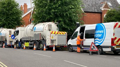 A white branded Thames Water van parked behind two large water tanker trucks. There are orange cones and road signs advising drivers of the disruption. A utility worker wearing orange hi-vis is closing the sliding door of the van. In the background there are trees and red brick houses with slate roofs. 