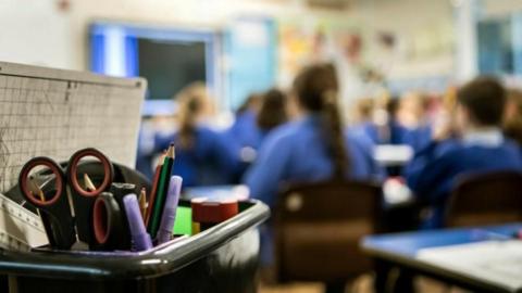 A pot full of stationary, including scissors, rulers and pencils, in the foreground as school children wearing blue jumpers face the opposite way in a classroom.