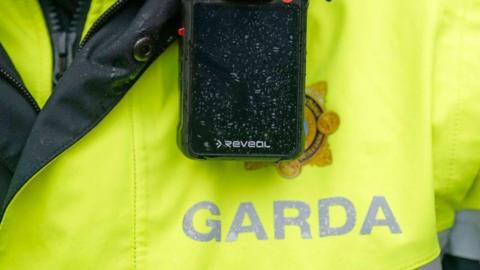 A radio is seen attached to an Irish police officers high viz jacket, marked with the word Garda
