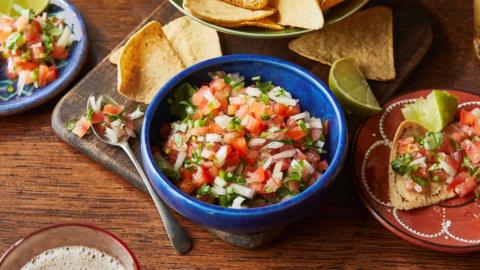 Diced tomatoes, green chilli, onion and coriander in a blue bowl next to a plate of tortilla chips