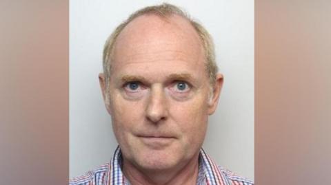 Mugshot of Christopher Cave. He has short blonde grey hair. He is wearing an open-necked, checked shirt.