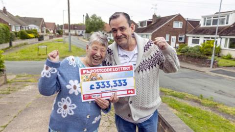 A couple hold up a cheque for £333,333 with their fists in the air. Nancy is wearing a light blue cardigan with white flowers across it. Colin is wearing a cream coloured cardigan with a maroon pattern on the upper section.
