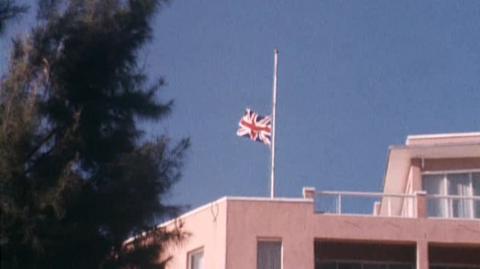 Union Jack flying at half mast on top of a pink coloured building with blue sky behind and a tree in the foreground on the left of frame.