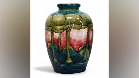 A small vase that has been painted with light green trees and dark green rolling hills. The sky has been painted a patchy pink and red colour.