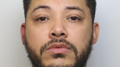 A man staring straight ahead with a blank expression. He has short, dark hair and a short beard. 