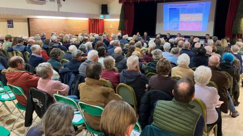 A large audience sits in rows of chairs inside a hall, facing a stage where several panellists are seated beneath a projected presentation.