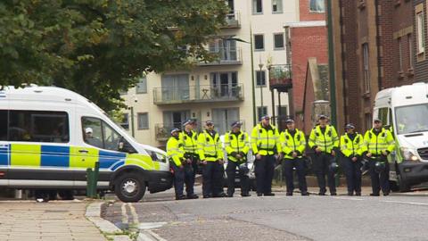 A row of police officers in high-viz jackets block off an empty street, two police vans either side of them.
