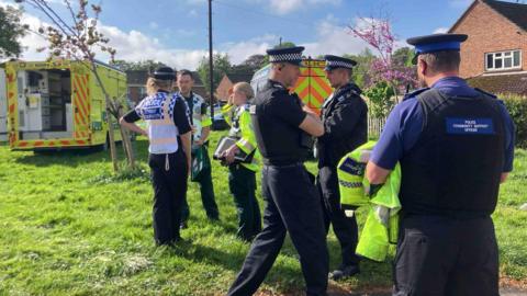 PCSOs, police officers and paramedics are standing on an area of grass in a residential area chatting. 
