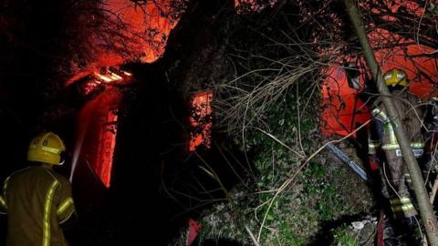 Backs of two firefighters with stone building in the background - it is engulfed by fire that is giving off a bright orange glow.