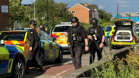 Armed officers walking down the street with police cars parked on the road