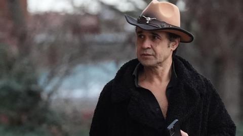 Chico arriving at court, wearing a brown cowboy hat with a silver cattle head adornment. He wears a black furry coat with no shirt on underneath and holds a mobile phone.