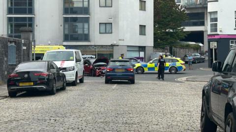 Several Devon and Cornwall Police officers patrol the scene of a crash involving a motorbike and car. The motorbike cannot be seen but the red Citreon DS3 involved in the incident is on the road with significant damage to its front. A police car is parked on the road along with an ambulance.
