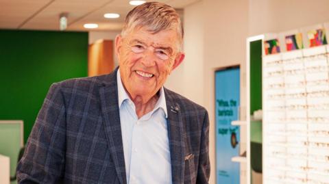 Man in a jacket which is blue and white and a light blue shirt. He has grey and white hair and is in a Specsavers store