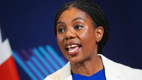 Kemi Badenoch is wearing a white jacket and a blue top. She is speaking in front of a blue background.