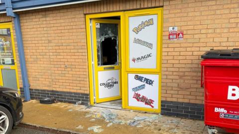 A shop front with a yellow door with a smashed glass window.