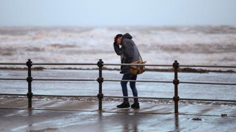 A man clutches his hat while walking along a windy seafront with rough waves in the background