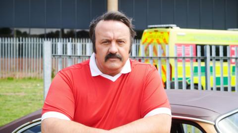 Mike Bubbins mewn crys coch a gwyn yn yr awyr agored gydag adeilad, ffens ac ambiwlans yn y cefndir