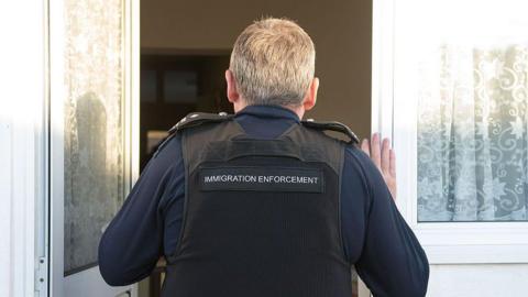 The back of a man with grey hair opens a front door - he is wearing a navy blue long sleeved top and a black vest which reads "Immigration Enforcement"