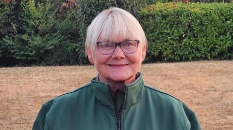 A woman with short blonde hair and glasses looking at the camera. She is wearing a green top with a zip. Light brown grass and a hedge are in the background.