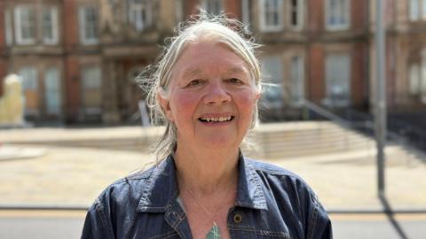 Joan Walley, a woman wearing a denim jacket, is standing outside Hanley Town Hall. She is smiling for the photograph.