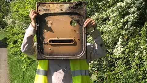 A litter womble holds up a rusty piece of metal resembling a face in front of her