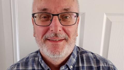 Andy Sturrock is stood against a door and has a short grey beard and dark framed glasses.