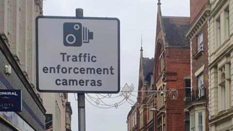 A Traffic enforcement camera sign, in a town centre, with stone and brick buildings, on either side of the sign. There are lights across the road. 