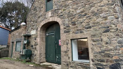 A stone wall building, with grey and brown, with a two windows and a large green arched doorway. The door has writing on it which says 'Sark Electricity Limited'. 