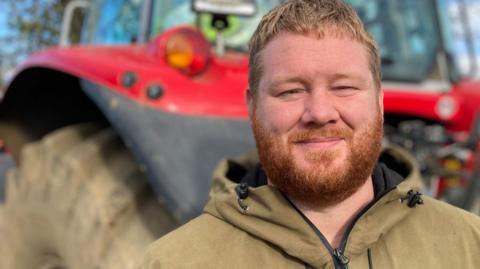 Man looking at the camera wearing a khaki green hooded jacket with a beard. He stands in front of a large red tractor