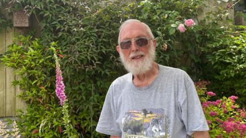 A man with grey hair and glasses with a grey tshirt standing in a garden with pink flowers and a wooden fence