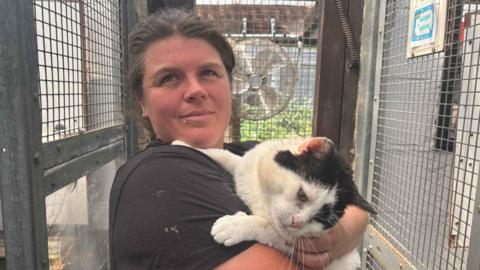 Emma Floyd is standing just off centre wearing a black T-shirt looking at the camera. She is holding a white and black cat in her arms, and is standing in a metal animal cage.