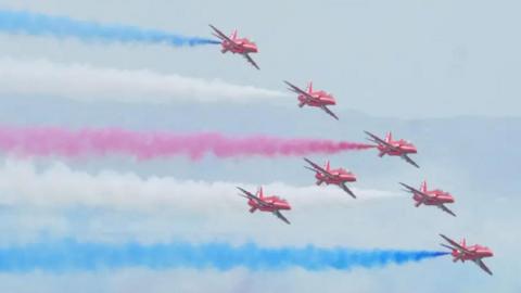 Aerial shot of seven Red Arrow jets in the sky from Airbourne 2024  in formation with red, white and blue smoke coming out of the rear of the planes.  