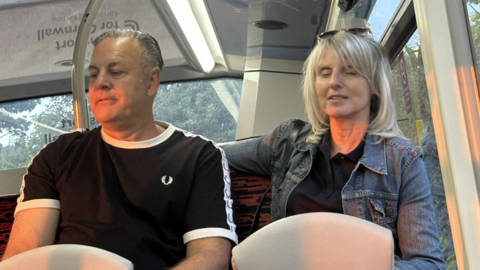 A man and a woman are pictured sitting on the back seat of a bus. The middle-aged man has slicked-back grey hair and is wearing a black Fred Perry T-shirt with a white trim. The woman is slim and has mid-length blonde hair and she is wearing a denim jacket. She has sunglasses on the top of her head.
