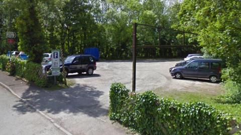 Street view of the car park in Rowsley. It has hedges at the front and trees in the background.