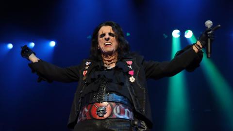 Rock star Alice Cooper on stage with his arms thrown in the air, a microphone held in one hand. He has black hair, heavy black eye makeup, black clothes, and a thick red belt with a silver skull on it.