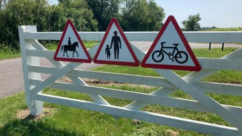 A white gate has four triangular road signs. The signs ask people to be aware of horse riders, people walking and cyclists.