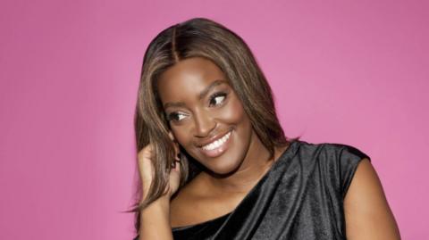 Woman wearing a black dress looking away from the camera to her right during a photoshoot. She is wearing make up and the wall / background behind her is purpley-pink