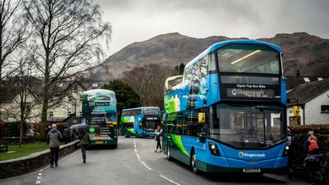 Three blue Stagecoach double decker buses on a village street in Cumbria with the Lake District fells in the background. The bus in the foreground is open-topped towards the rear. It is on the right of the road - which is flanked by a smattering of white buildings - with another standard bus a little way behind it. The third bus is on the left hand side and is seen from the rear. It is a 599 service. There are people queuing for the open-topped bus, some getting on the 599, a person crossing the road and two walking towards the bus on the left. One has an umbrella raised.