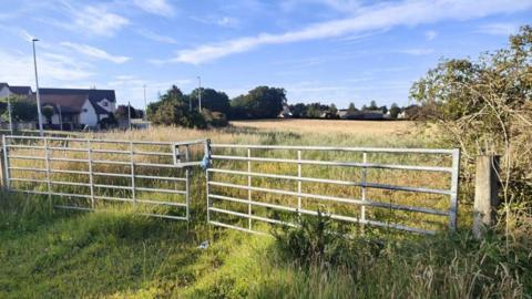The sun is glinting off a metal gate that encloses a large field. Houses can be seen around the edge and there are bushes and trees.