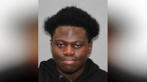 Umar Dumbuya is looking at the camera with a slight toothless smile. He is wearing a black hoodie with the hood down. he has a medium afro on top and shaved at the sides
