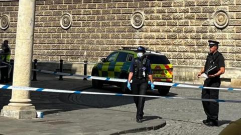 Police officers wearing black uniform and blue gloves, in front of a blue and white police tape, with police car in distance.