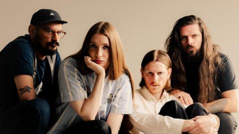 The band Dry Cleaning. There are three men and one woman lined up next to each other. Three of them are crouching, and one is sat on the floor with their knees up to their chest.  