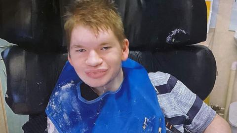 Declan Horton. He has short brown hair and is sat in a black wheelchair. He is wearing a blue bib over a striped t-shirt that has some food residue on it. He is looking at the camera and smiling.