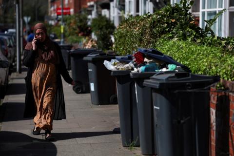 A woman holds a phone up to her ear as she walks towards us on a residential street, past some black bins - two of which are overflowing.
